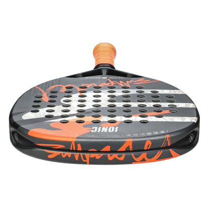 A close-up of the Bullpadel Ionic Power 2025 Padel Racket, with a textured grey and orange surface featuring holes for the XFORCE System. The orange grip complements white and orange face markings.