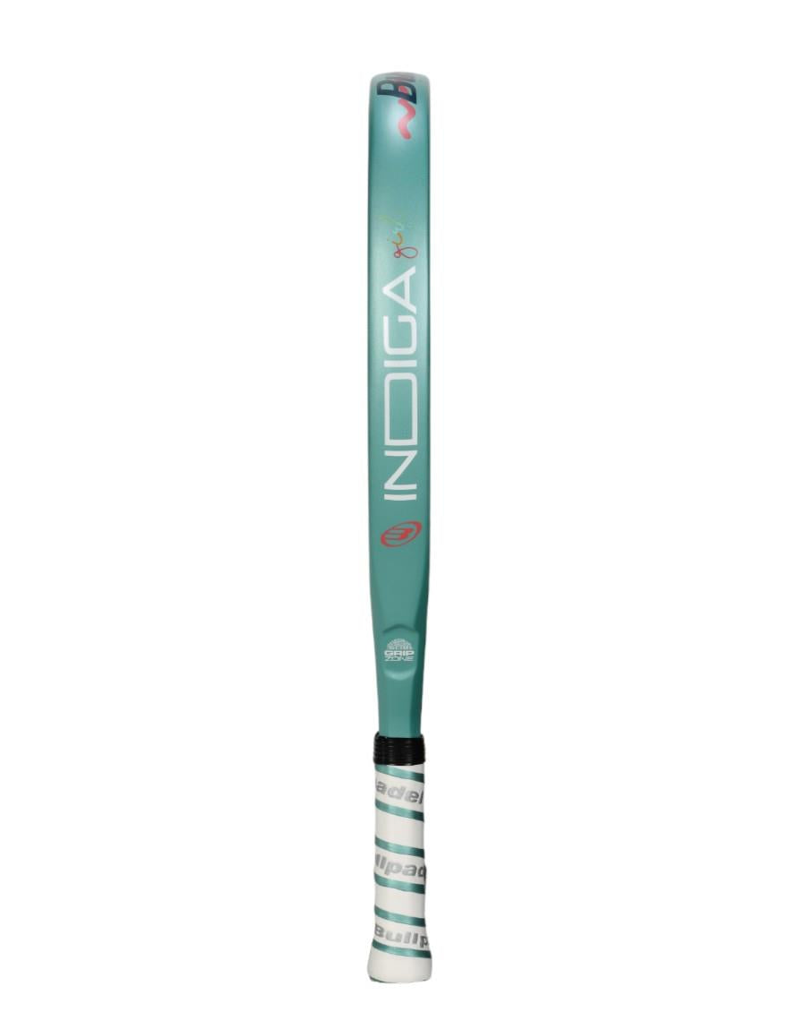 A teal Bullpadel Indiga Junior 25 Girls padel racket features a black-accented body with "Bullpadel" inscribed in white. The handle sports Grip Zone technology and is wrapped in a white grip with grayish-green patterns.