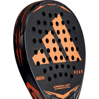 A close-up of the adidas Arrow Hit Carbon CTRL 2026 padel racket shows a large orange "A" logo, multiple round holes on the face, and "ARROW-HIT CARBON CTRL" text near the bottom.