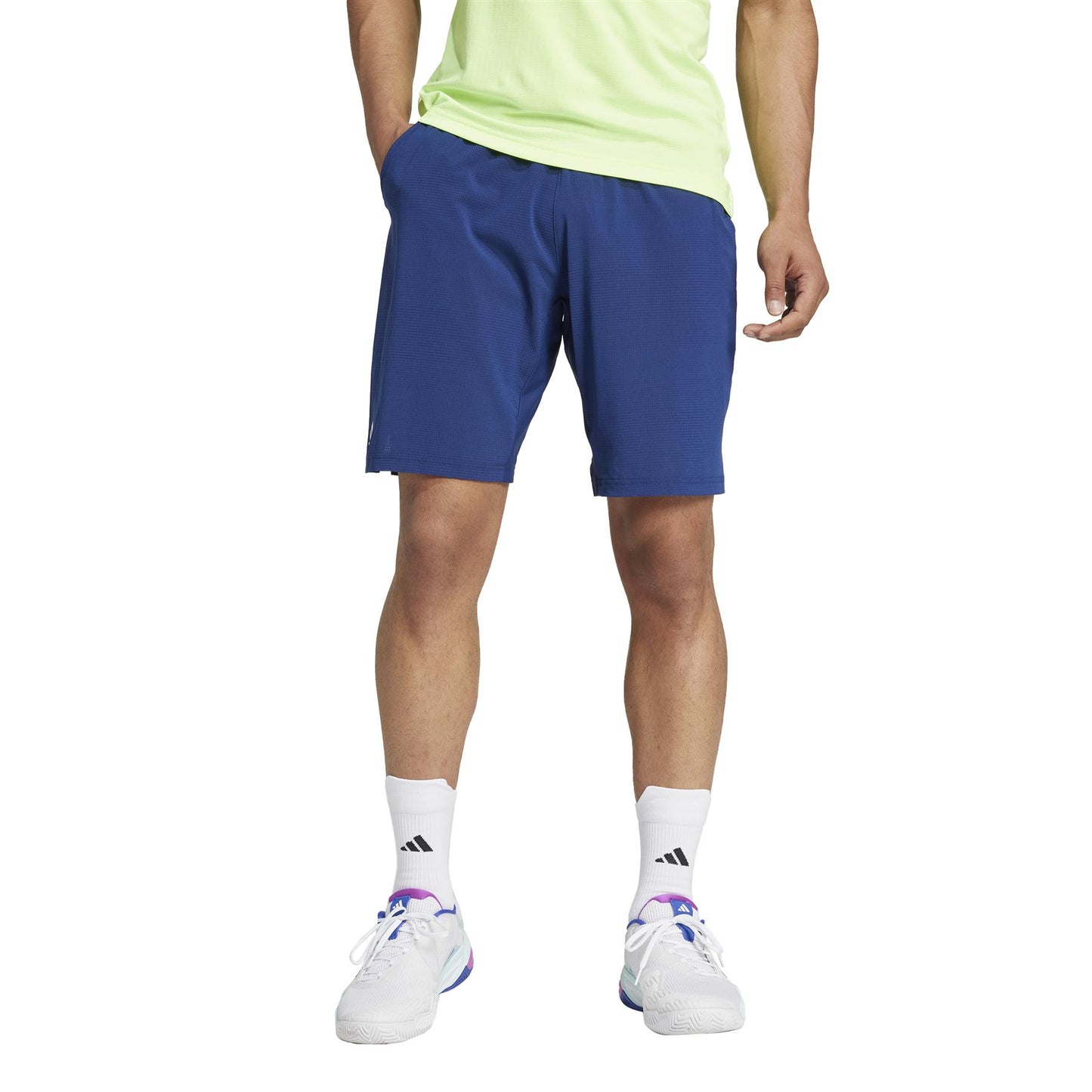 A person stands with hands in pockets against a white background, wearing a light green T-shirt, navy ADIDAS Ergo Men's Padel Shorts by adidas, white Adidas socks, and white sneakers with purple accents.