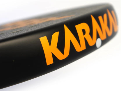 Close-up of the edge of the Karakal Air Lite 345 Orange Padel Racket, featuring bold orange "Karakal" branding in focus against a blurred background.