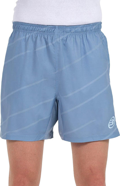 A person wears Bullpadel Cabanas Mens Padel Shorts in shadow blue, featuring a subtle diagonal stripe pattern, white logo on the lower left leg, Quickerdry technology, and an elastic waistband. A white shirt is partially visible.