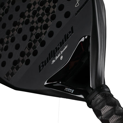 A close-up shot of the Bullpadel Icon 2027 Padel Racket displays its textured black surface, iconic Bullpadel logo, and a black-gripped handle. The design showcases X-Tend Carbon 12K detailing, “Icon Core” branding, and features an advanced MultiEva core from Bullpadel for superior performance.