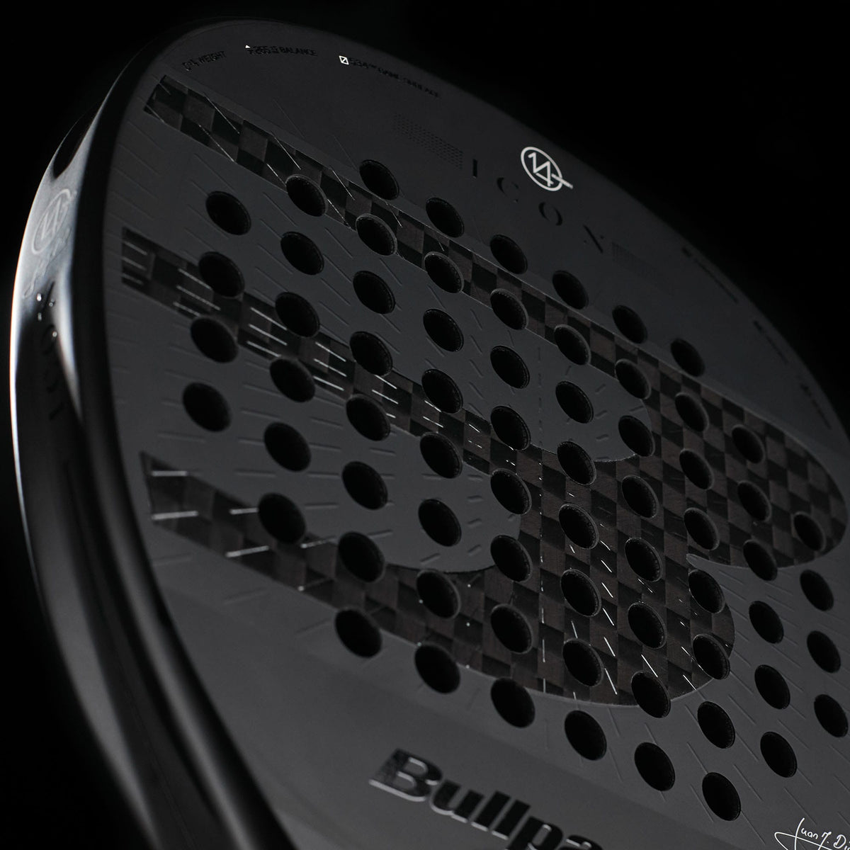 A close-up of the Bullpadel Icon 2027 Padel Racket showcases its dark design with round perforations, highlighted by a subtle bull motif and the Bullpadel logo against a black backdrop. Engineered with X-Tend Carbon 12K construction and a MultiEva core, this racket delivers enhanced performance.