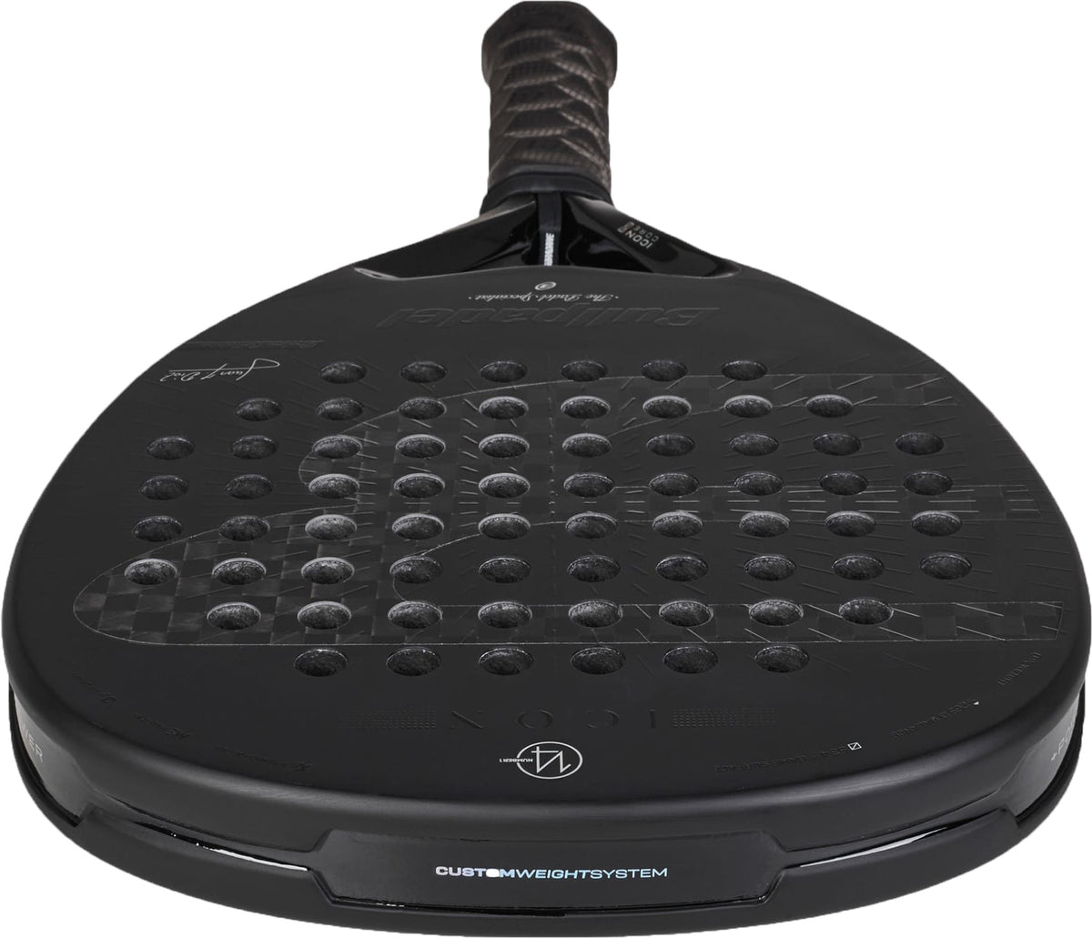 A close-up view of a black Bullpadel Icon 2027 Padel Racket, showcasing its textured X-Tend Carbon 12K surface with multiple circular holes, padded handle, MultiEva core, and Bullpadel branding details.