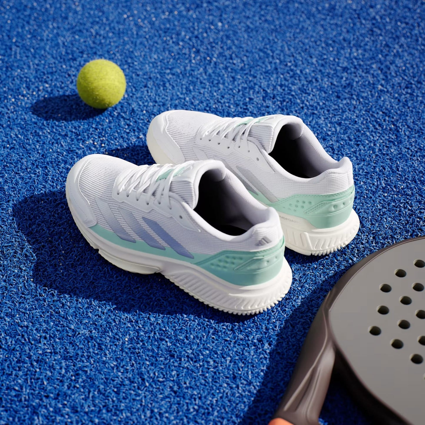A pair of adidas Courtquick Women's Padel Shoes in Cloud White, Silver Metallic, and Clear Mint rests on a bright blue tennis court beside a yellow tennis ball and a black paddle racket.