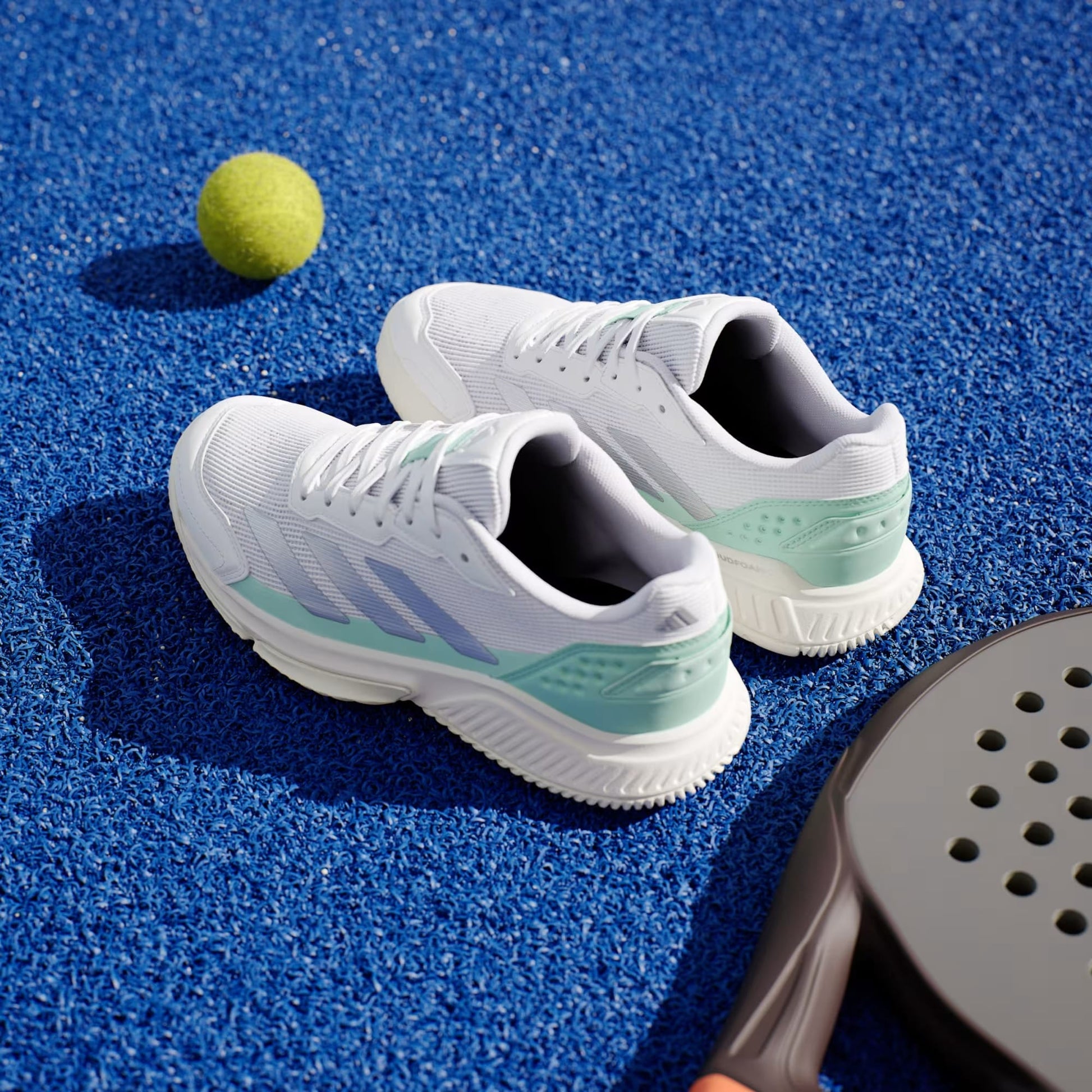 A pair of adidas Courtquick Women's Padel Shoes in Cloud White, Silver Metallic, and Clear Mint rests on a bright blue tennis court beside a yellow tennis ball and a black paddle racket.
