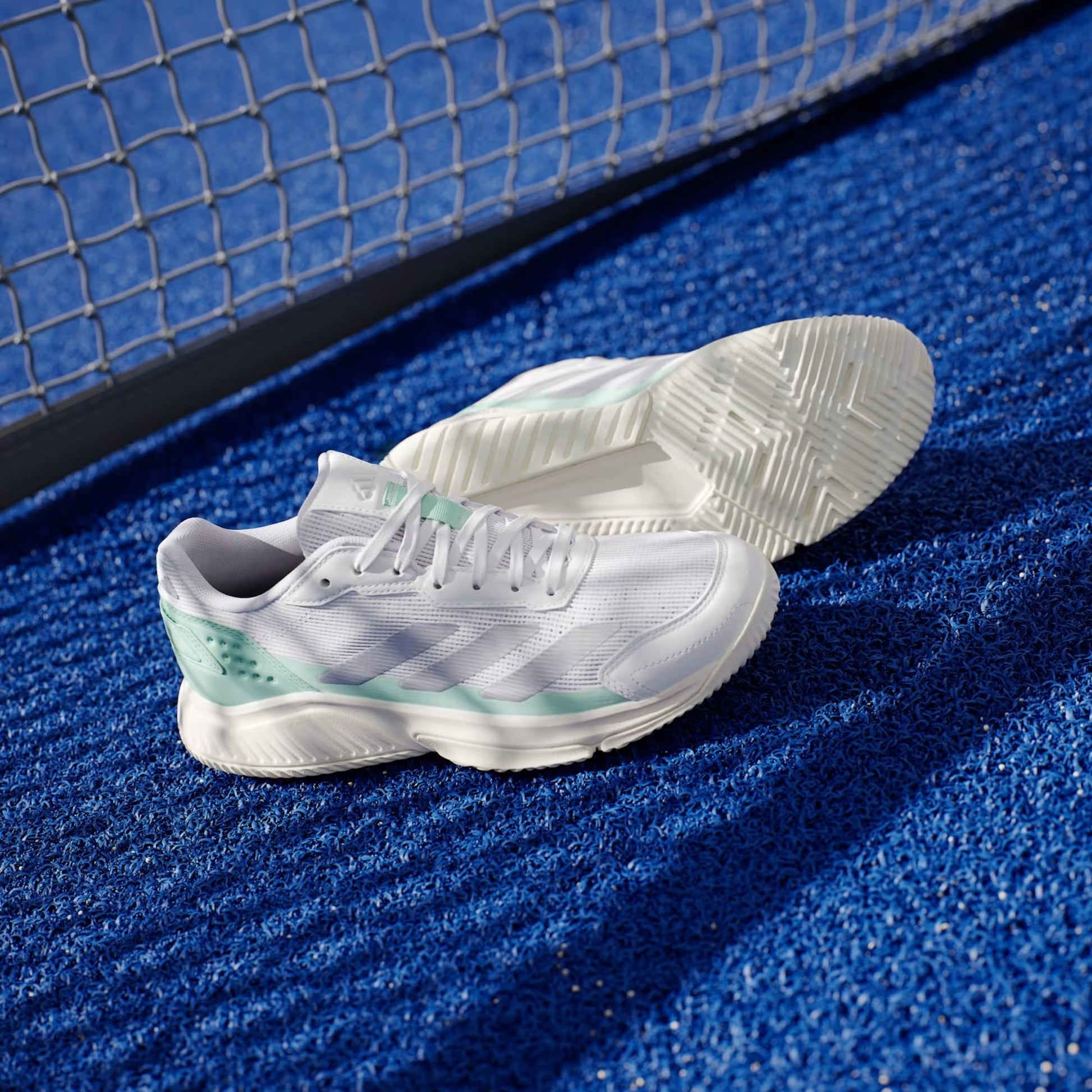 A pair of adidas Courtquick Womens Padel Shoes in Cloud White, Silver Metallic, and Clear Mint rest on a bright blue textured court near a metal fence.