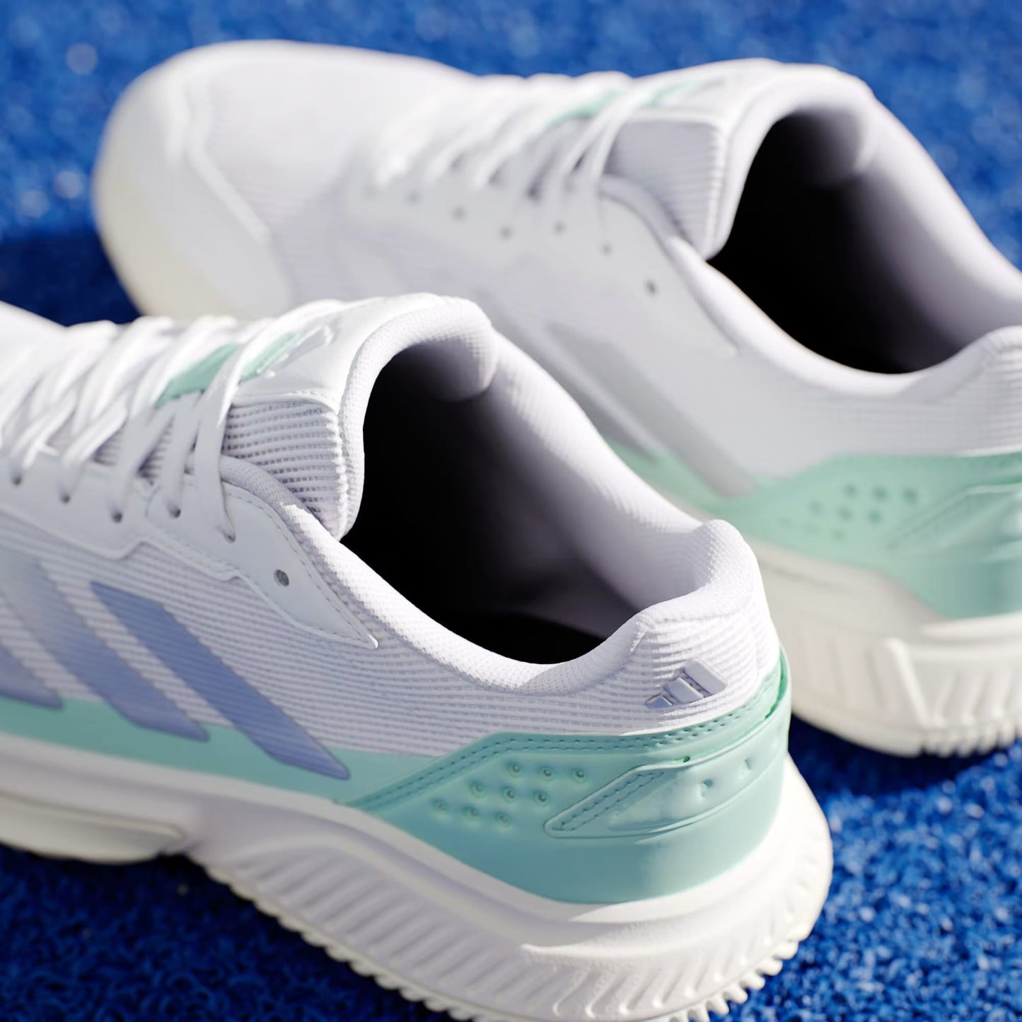 A close-up of adidas ADIDAS Courtquick Women's Padel Shoes in Cloud White, Silver Metallic, and Clear Mint displayed on a bright blue textured surface.