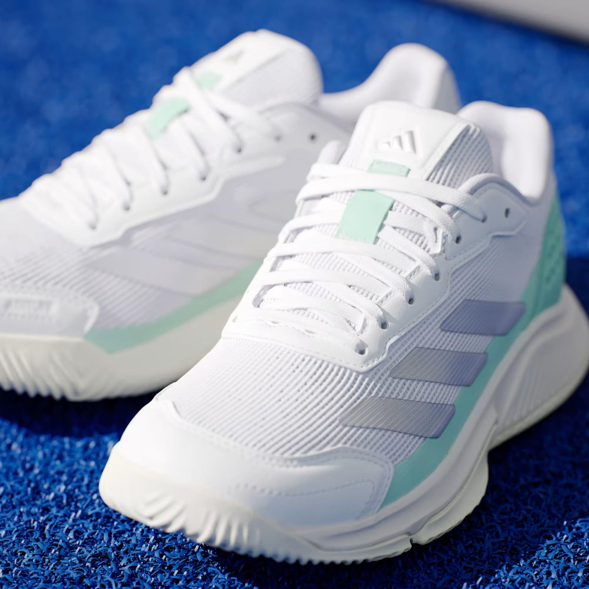 A close-up of adidas Courtquick Womens Padel Shoes in Cloud White/Silver Metallic/Clear Mint, with laces and the adidas logo, set on a textured blue surface—ideal for stylish padel enthusiasts.