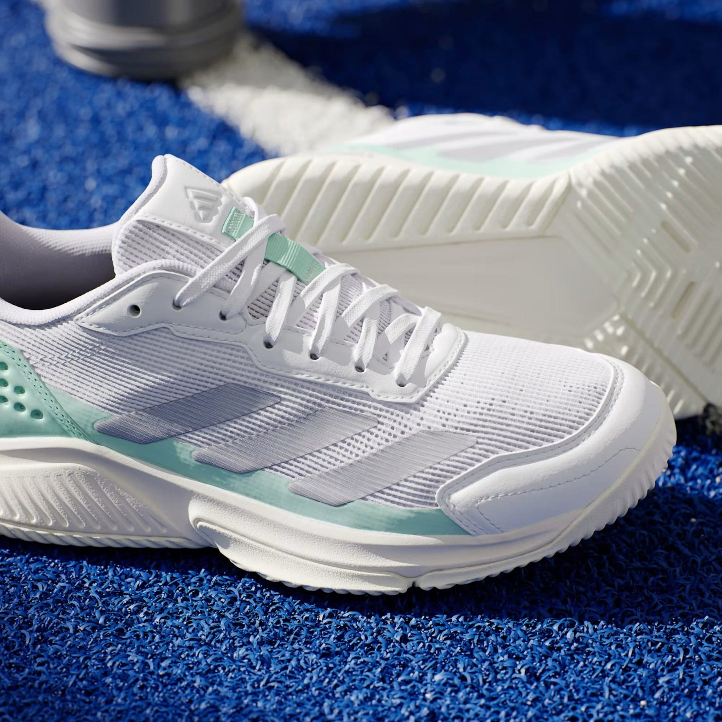 A close-up of adidas Courtquick Women's Padel Shoes in Cloud White, Silver Metallic, and Clear Mint on a blue textured court with a white line—perfect for those seeking stylish women's court shoes.